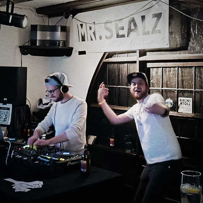 Flo mixing behind the decks while Dave dances beside him, under a Mr. Sealz banner.